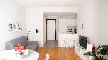 Un appartement moderne comprenant un salon, une cuisine et un coin repas. On y trouve un canapé gris, une table basse blanche et un téléviseur. La cuisine est équipée de meubles blancs et d’un plan de travail.