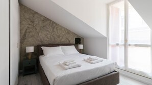 Un dormitorio con techo inclinado, una cama con ropa blanca, dos mesitas de noche con lámparas y un ventanal con persianas.
