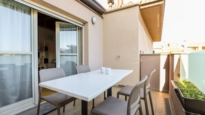 A balcony with a white table and four chairs, a sliding glass door, and a small garden area with plants. - Colombo 4 (Villasanta)