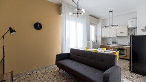 A modern kitchen with white cabinets, a black refrigerator, and a dining area with a gray sofa and yellow chairs. - Quarnaro 2 (Monza)
