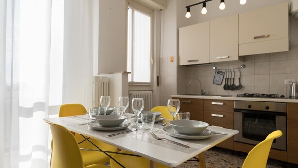 A modern kitchen with white cabinets, a dining table set with plates, glasses, and cutlery, and yellow chairs. - Quarnaro 2 (Monza)