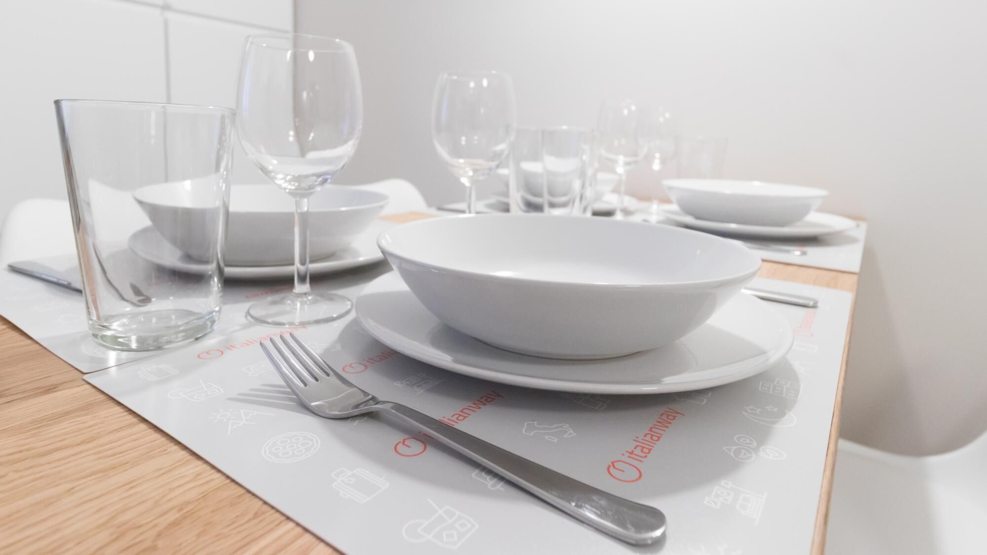 Una mesa dispuesta con vajilla blanca, incluyendo cuencos y copas de vino, sobre un mantel individual con iconos.