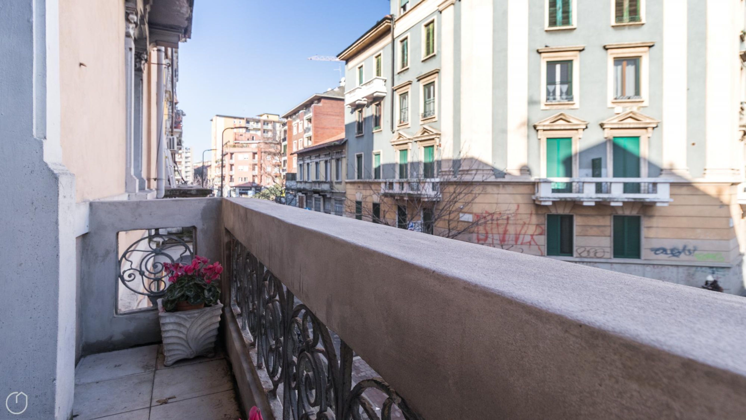 Un balcon orné d’une balustrade en fer forgé, avec un pot de fleurs et une vue sur les bâtiments voisins.