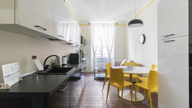 A modern kitchen with a sink, kettle, toaster, and coffee maker. There is a dining area with yellow chairs and a table. - Italianway Easy - Buenos Aires 65 B (Milan)