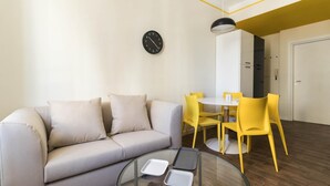 A modern living room with a sofa, a round glass coffee table, and a dining area with yellow chairs and a white table. - Italianway Easy - Buenos Aires 65 B (Milan)