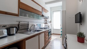 Cocina moderna con horno empotrado, hervidor y cafetera. Hay una ventana que proporciona luz natural y un televisor montado en la pared.