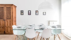 Un comedor con una mesa de vidrio dispuesta para seis personas, rodeada de sillas blancas. Hay cuadros enmarcados en la pared y un aparador de madera a la izquierda.