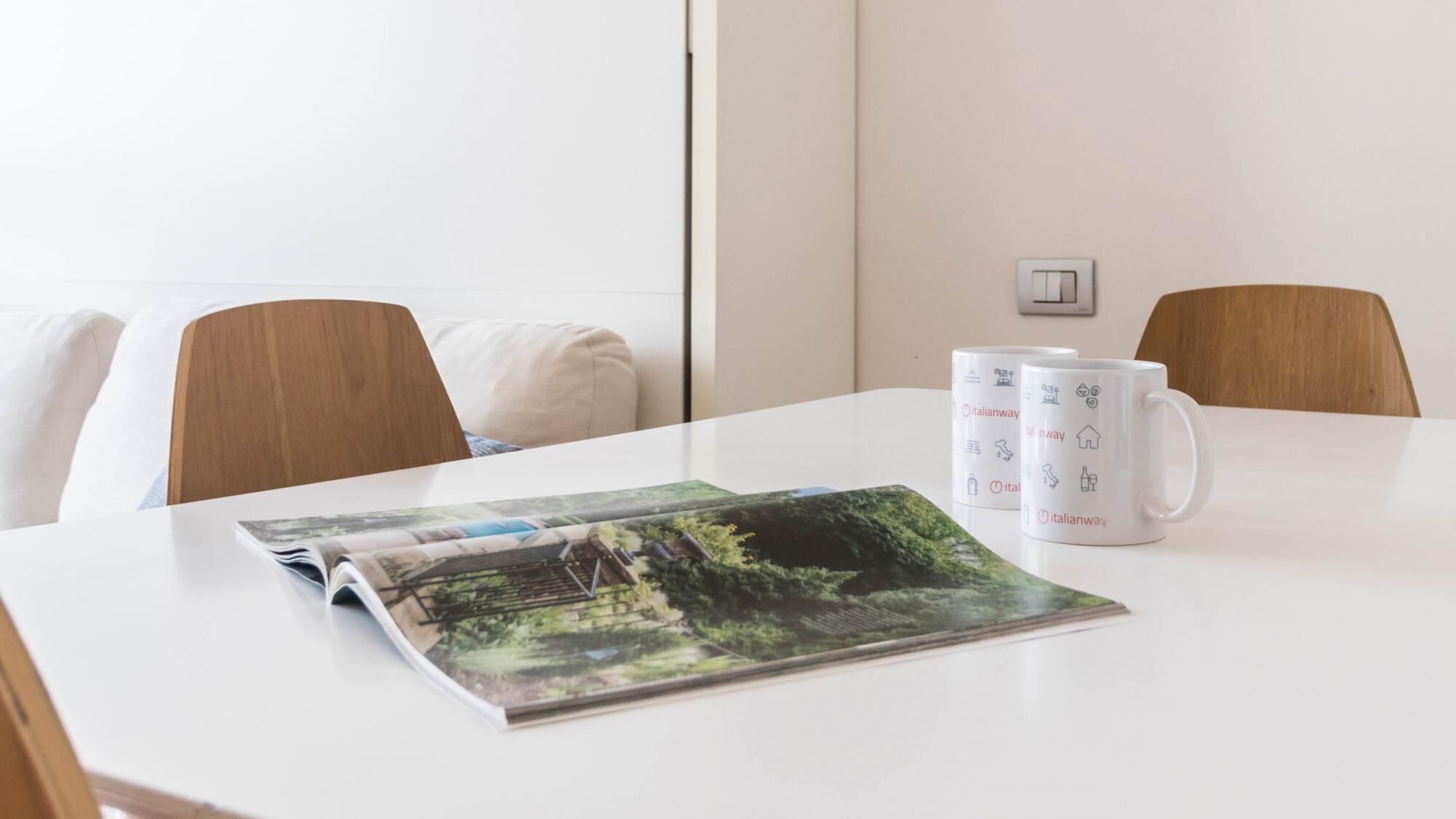 A white table with a magazine, two white mugs with Italianway branding, and a light switch on the wall.