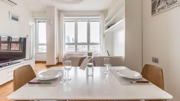 A modern dining area with a white table set for two, a television on the left, and a city view through large windows. - Italianway Easy - Biancamano 2 (Milan)
