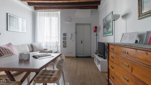 Ett modernt vardagsrum med ett matbord i trä, en soffa och en tv. I bakgrunden syns en vit dörr och en bokhylla fylld med böcker.