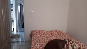 Un dormitori amb un llit, un coixí i una taula de nit. Hi ha una porta que dona a una altra habitació.