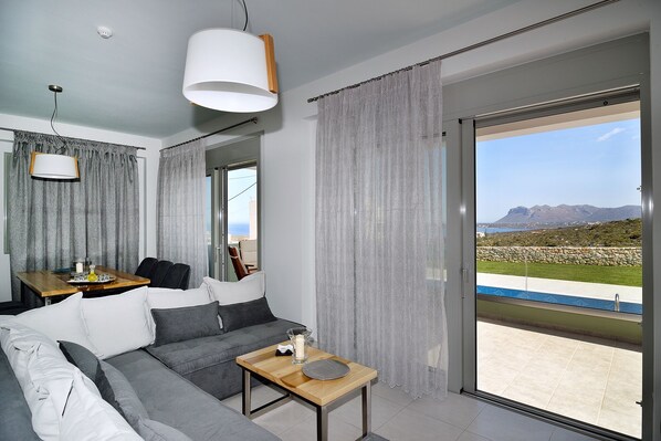 A modern living room with a grey sofa, a wooden coffee table, and a view of the sea through large sliding glass doors.