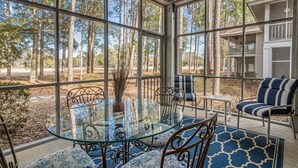 A sunroom with a glass table and chairs, surrounded by large windows offering a view of trees and a building.
