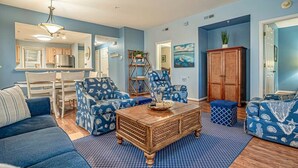 A living room with a blue sofa, matching armchairs, a wooden coffee table, and a kitchen in the background.