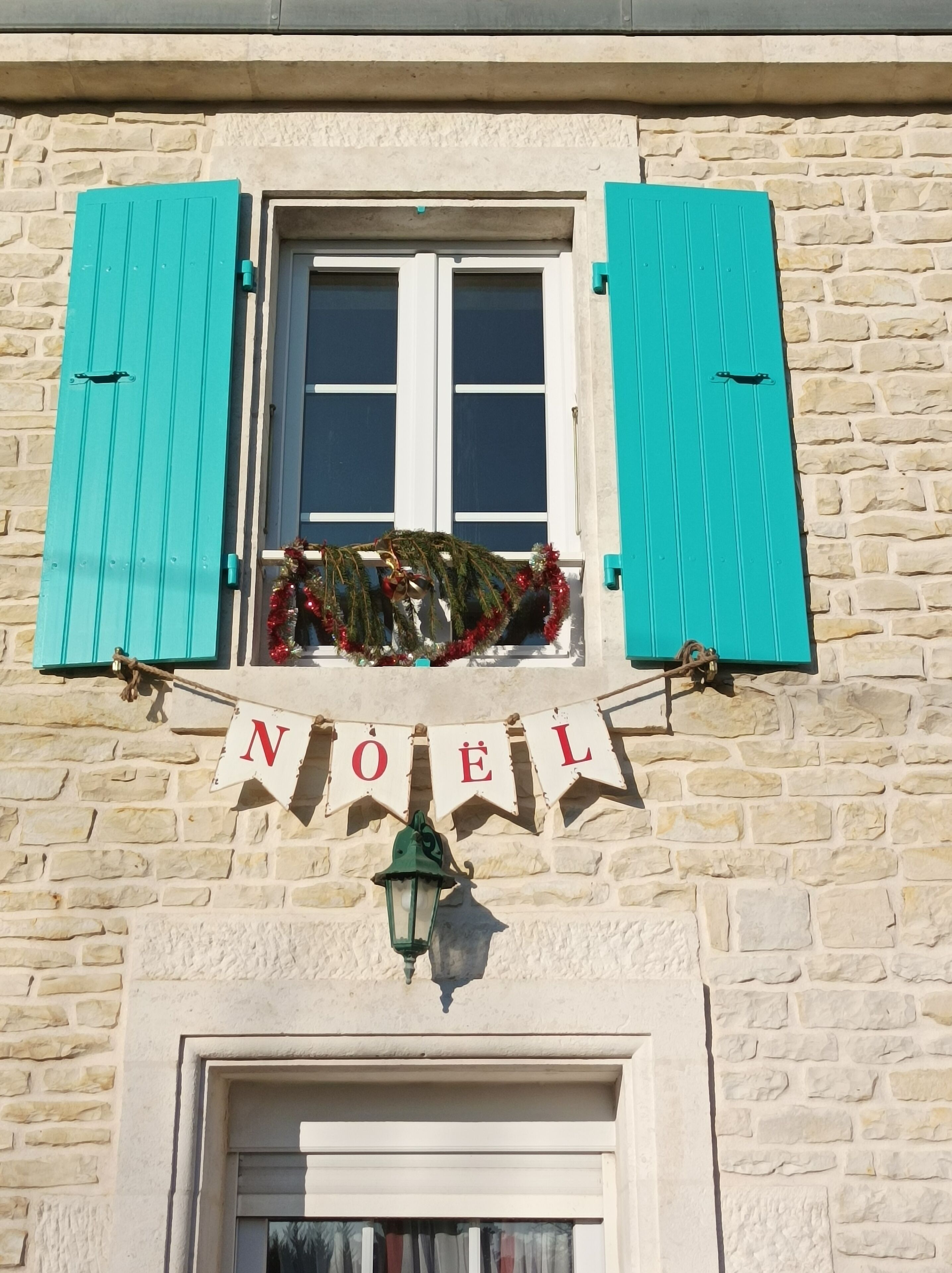 Ein Gebäude mit Steinwänden, einem Fenster mit türkisblauen Fensterläden, einem 'NOËL'-Schild und einem Laternen.
