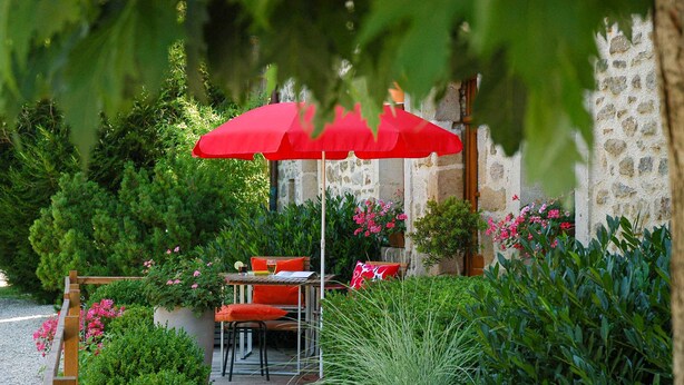 Una terraza de madera con mesa y sillas, rodeada de abundante vegetación y un muro de piedra.