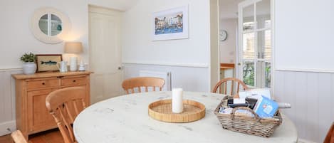 A dining area with a round table, wooden chairs, a white lamp, a framed picture, and a wicker basket with magazines.