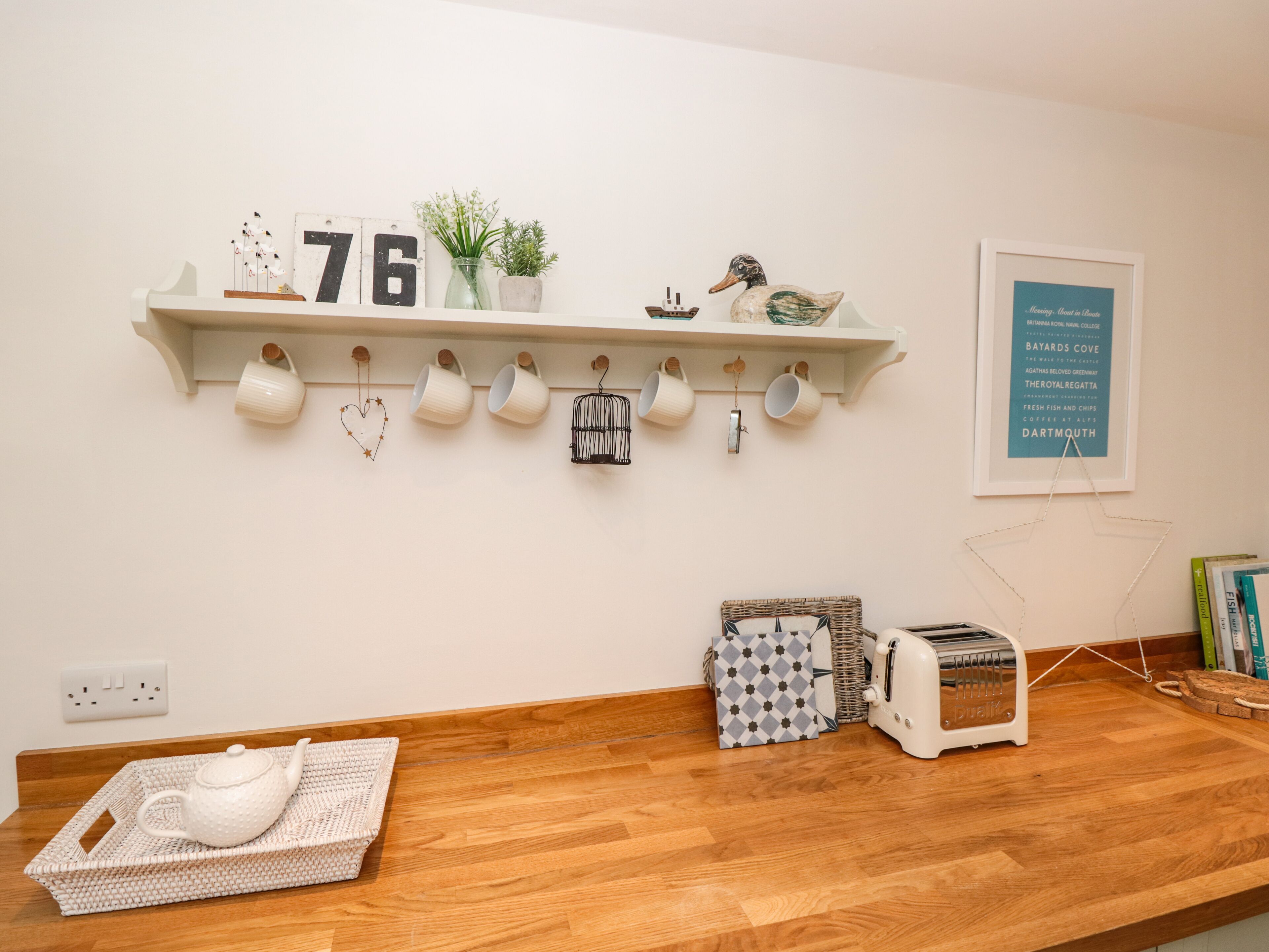A kitchen with a wooden countertop, a white teapot on a woven tray, a white toaster, and a shelf with mugs and a decorative number '76'.