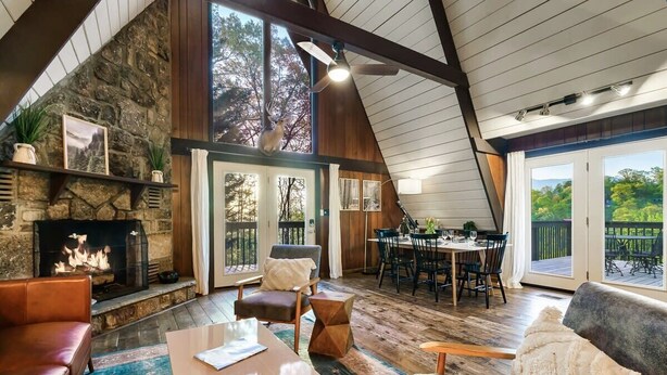 A cozy living room with a stone fireplace, wooden ceiling beams, and a large window overlooking a deck.