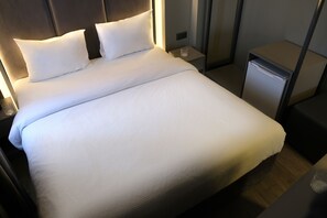 Een net opgemaakt bed met witte lakens en kussens, een nachtkastje met een glas, en een klein nachtkastje met een lamp.