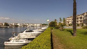 Uma marina com barcos atracados, um caminho bordeado por vegetação e um edifício com varandas ao fundo.