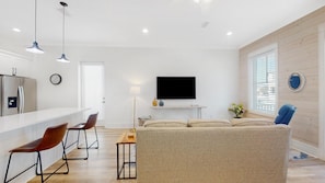 Un salon moderne avec un canapé, une table basse et un téléviseur fixé au mur. Il y a également un coin cuisine équipé d’un réfrigérateur et une table à manger avec des chaises.