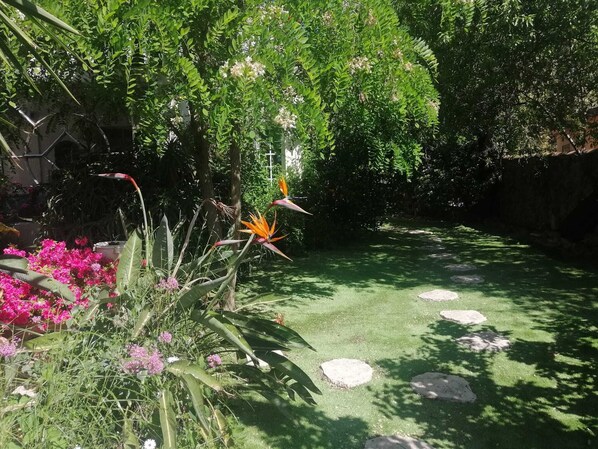 Un jardín con diversas plantas y flores, un sendero de piedra y una cruz al fondo.