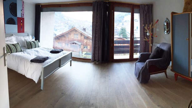 A bedroom with a bed, a chair, a wardrobe, and a view of a mountain town through a large window. - La Clusaz in a quiet location 300m from the village center (La Clusaz)