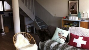 A living room with a staircase, a sofa with reindeer and Swiss flag pillows, a wicker chair, and a bookshelf with decorative items. - La Clusaz in a quiet location 300m from the village center (La Clusaz)