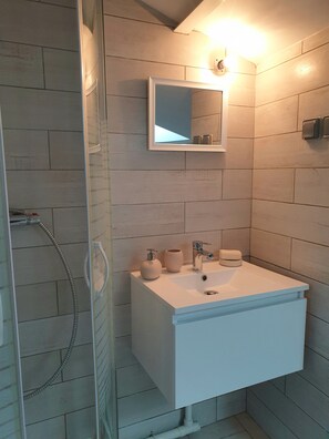 A bathroom with a glass shower enclosure, a white sink with a mirror above it, and a wall-mounted soap dispenser.