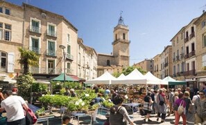 A bustling outdoor market with vendors and visitors, surrounded by historic buildings and a prominent tower.