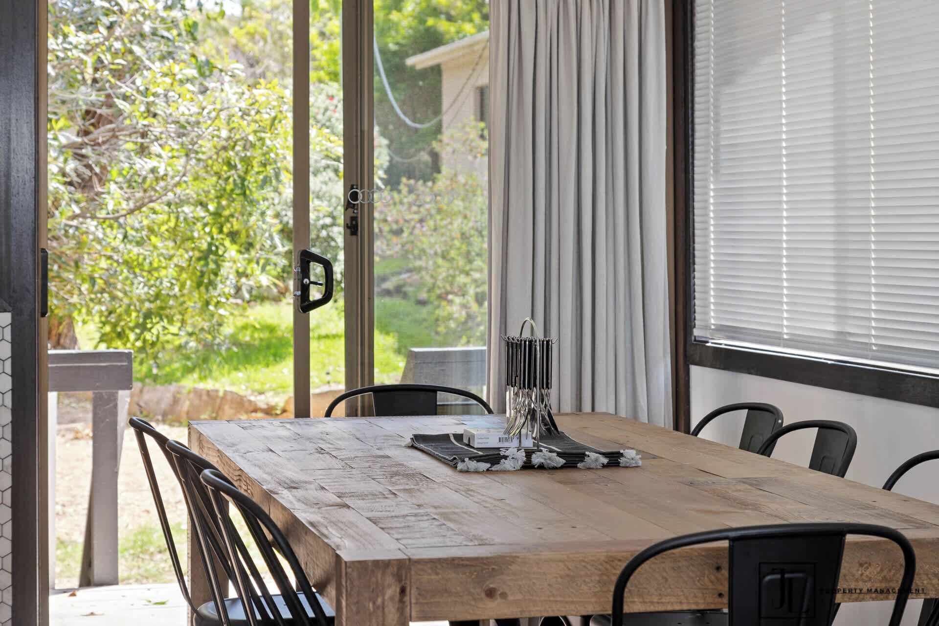 A wooden dining table with chairs, a window with blinds, and a view of greenery outside.