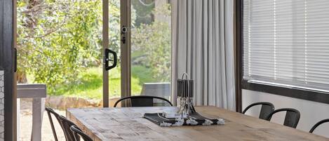 A wooden dining table with chairs, a window with blinds, and a view of greenery outside.