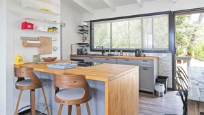 A modern kitchen with a wooden island, bar stools, a large window, and a view of greenery outside. - Waterfront Pet-Friendly Home - Oceanview Cottage (Malua Bay)