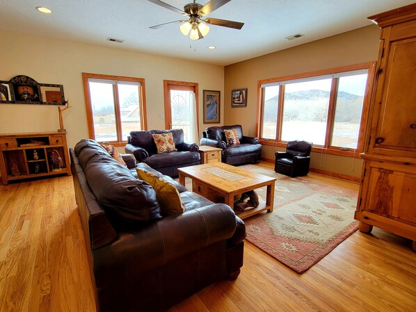 A spacious living room with leather sofas, a wooden coffee table, and large windows offering a view of the outdoors. - Casa La Capilla: Rural Creekside Retreat near SF (Santa Fe)