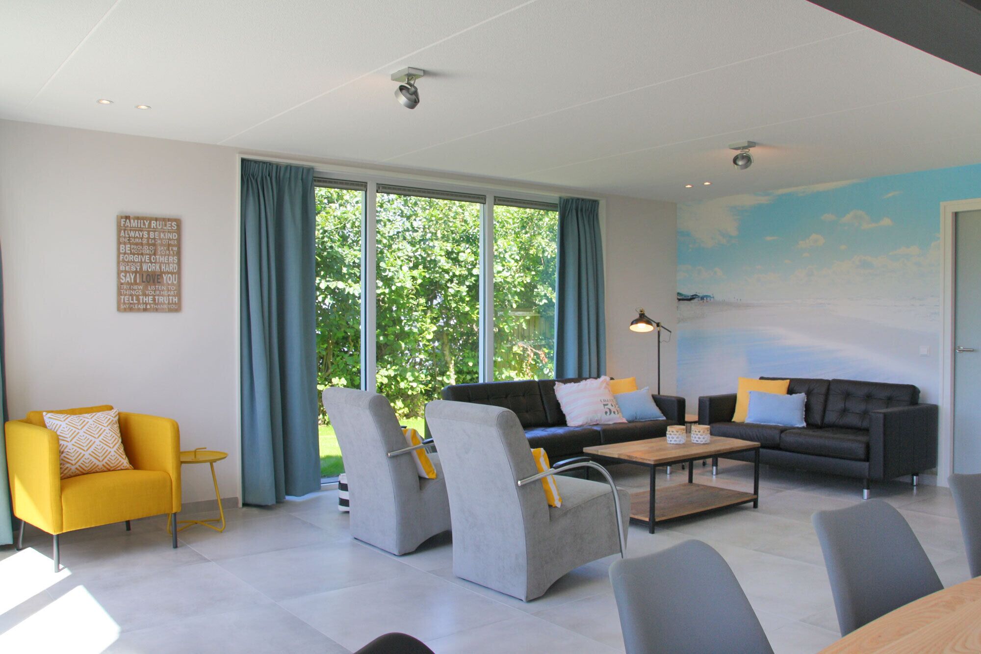 A modern living room with a sofa set, armchairs, and a coffee table. There are large windows with curtains, a wall-mounted light, and a ceiling-mounted camera.
