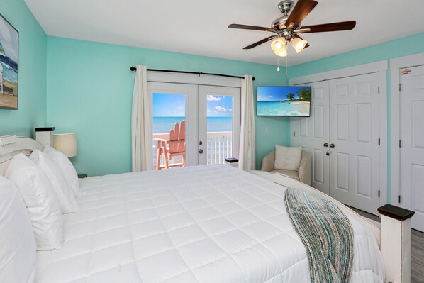 A bedroom with a large bed, a ceiling fan, a TV, and a door leading to a balcony with a view of the ocean.