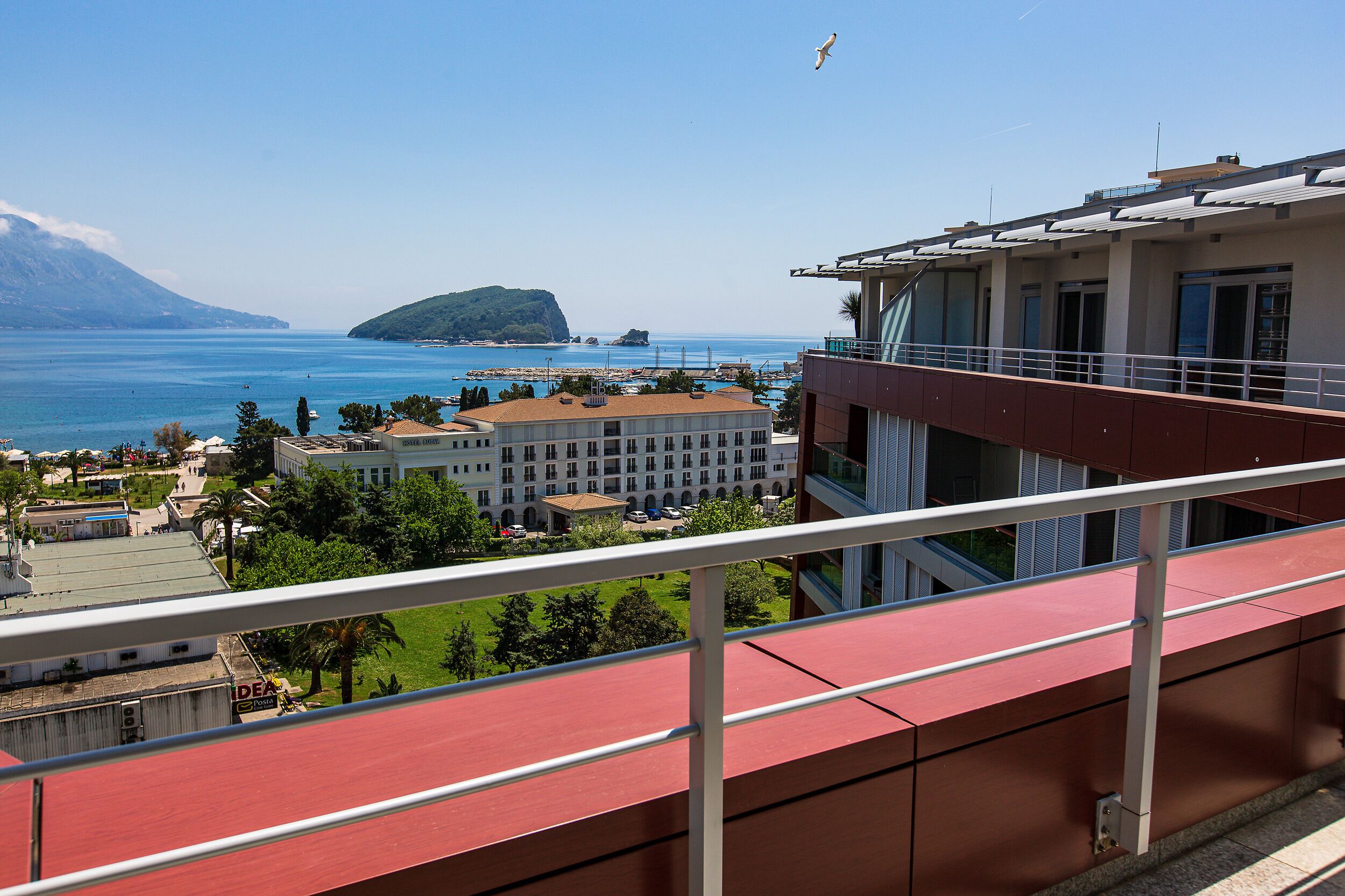 Un balcón con vistas a la costa, que incluye un edificio de techo rojo, una masa de agua y una isla distante.