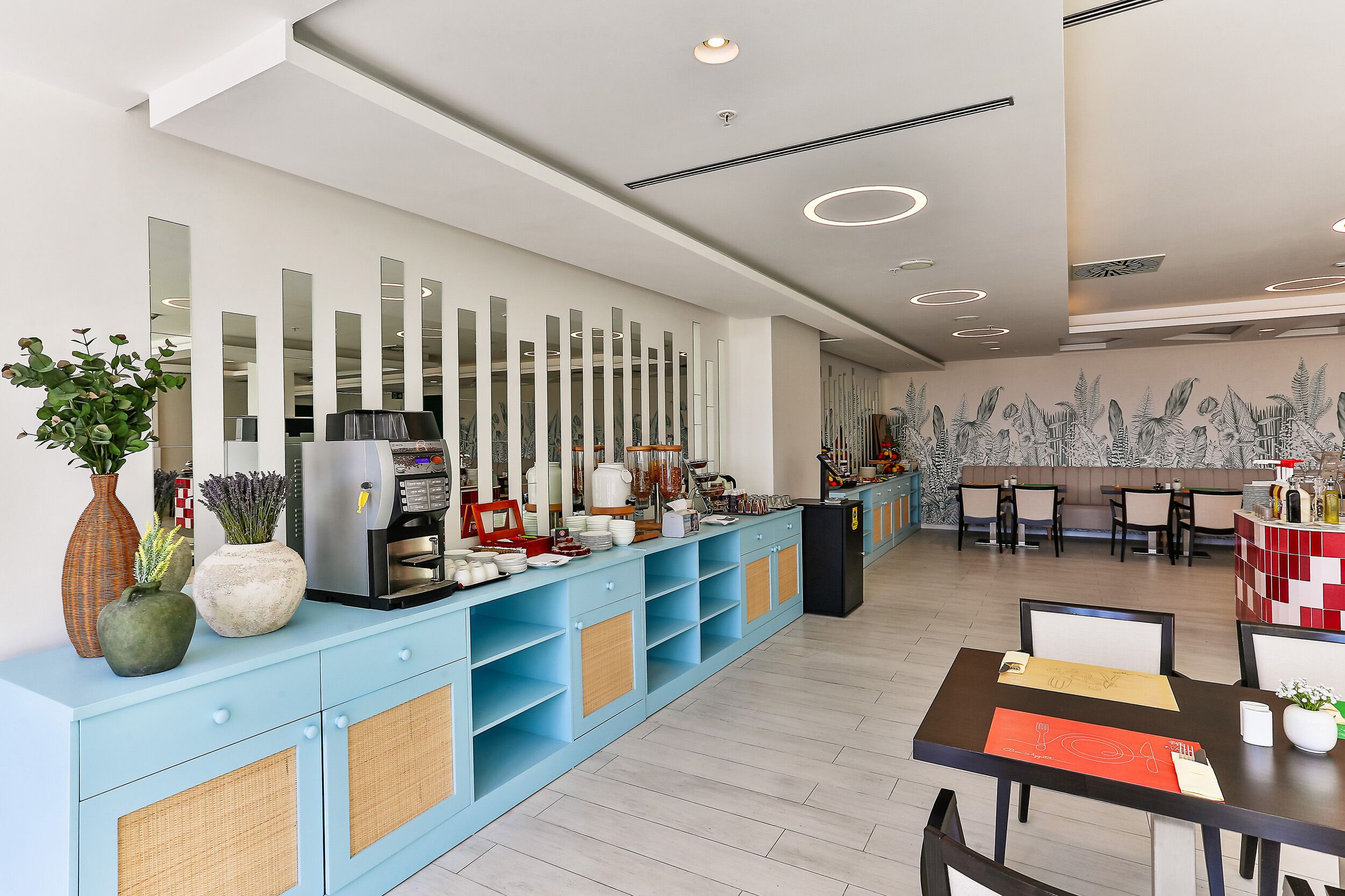 A modern hotel lobby with a light blue reception desk, a large mirror wall, and a dining area with tables and chairs.