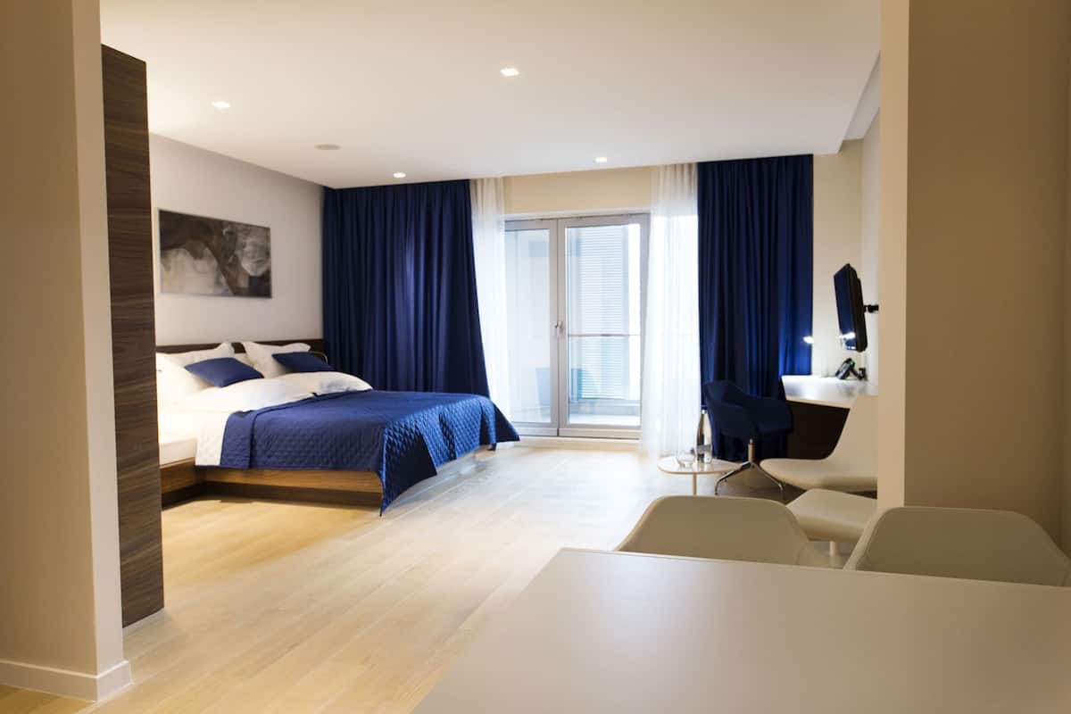 A modern hotel room with a large bed, a desk, and a chair. There is a window with curtains and a door leading to a balcony.