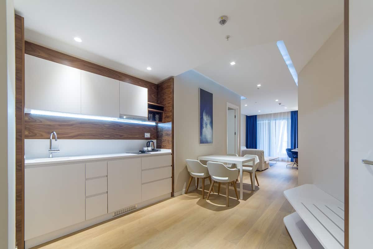 A modern kitchen with white cabinets, a stainless steel sink, and a built-in oven. There is a dining area with a white table and chairs, and a large window with blue curtains.