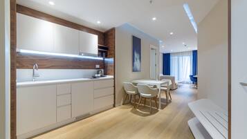 A modern kitchen with white cabinets, a stainless steel sink, and a built-in oven. There is a dining area with a white table and chairs, and a large window with blue curtains.