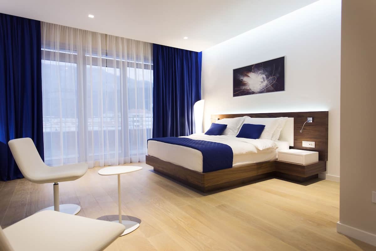 A modern hotel room with a large bed, a bedside table, a chair, and a view of the city through large windows.