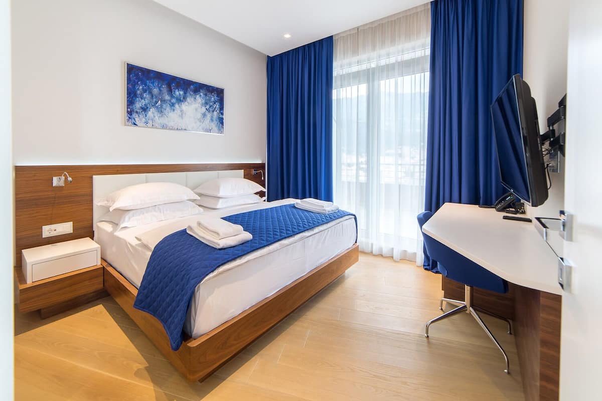 A modern hotel room with a wooden bed, a desk, a flat-screen TV, and a large window with blue curtains.