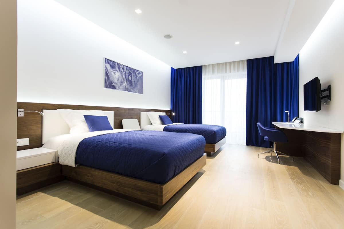 A modern hotel room with two beds, a desk, and a large window with blue curtains.