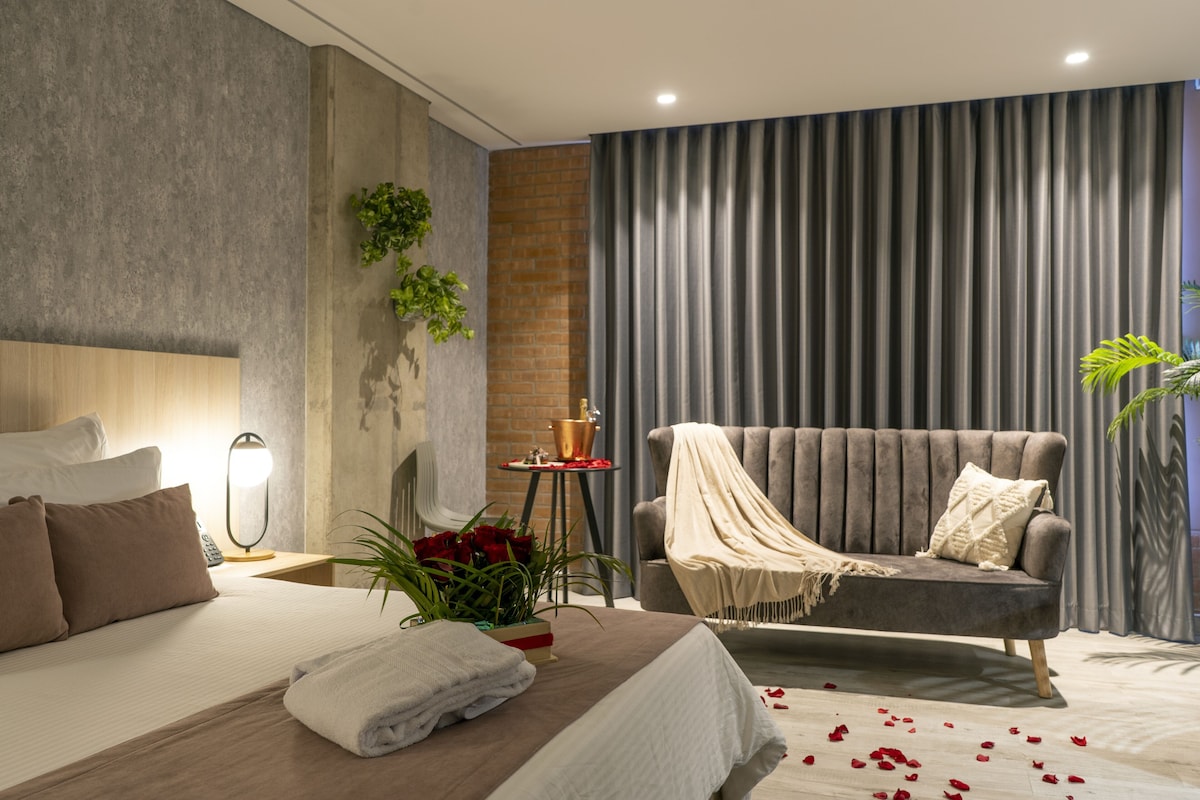 A modern hotel room with a large bed, a sofa, a small table with a lamp, and a potted plant.