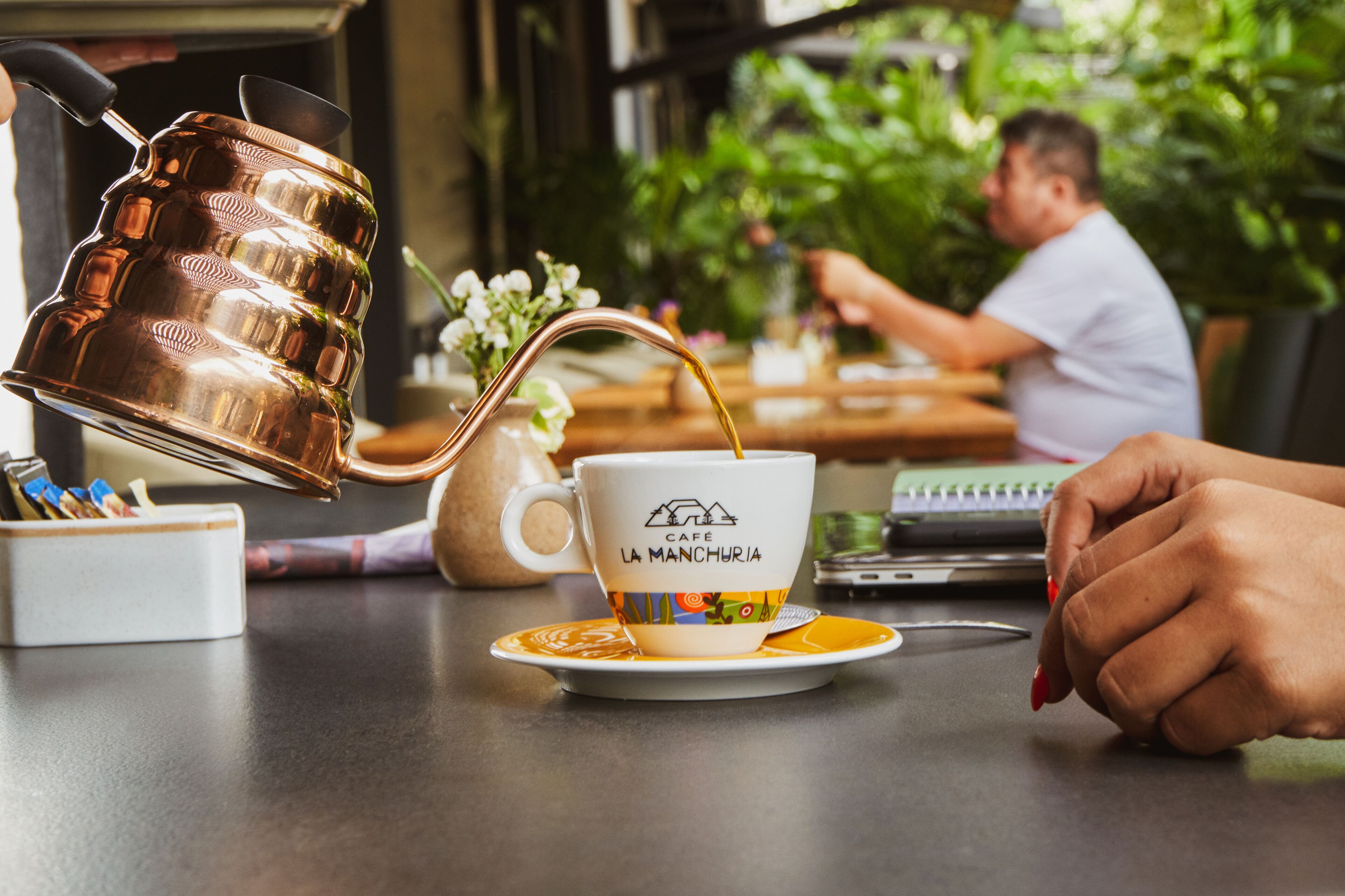 Una persona sirve café en una taza con la etiqueta 'Café La Manchuria' sobre una mesa con una computadora portátil y un cuaderno.