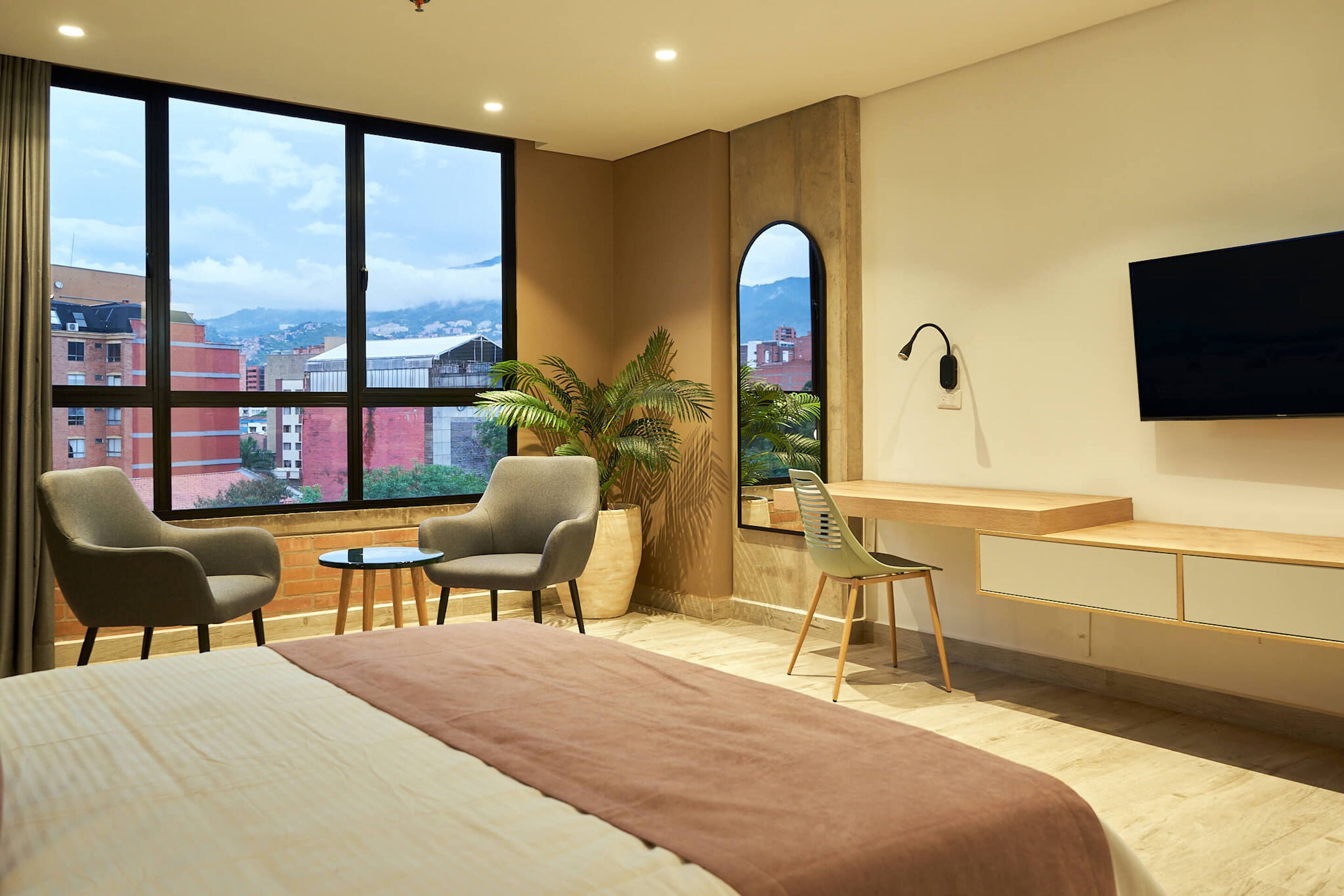 Ein Hotelzimmer mit einem großen Bett, zwei Sesseln, einem kleinen Tisch, einem Schreibtisch, einem Fernseher und einem großen Fenster mit Blick auf die Stadt.