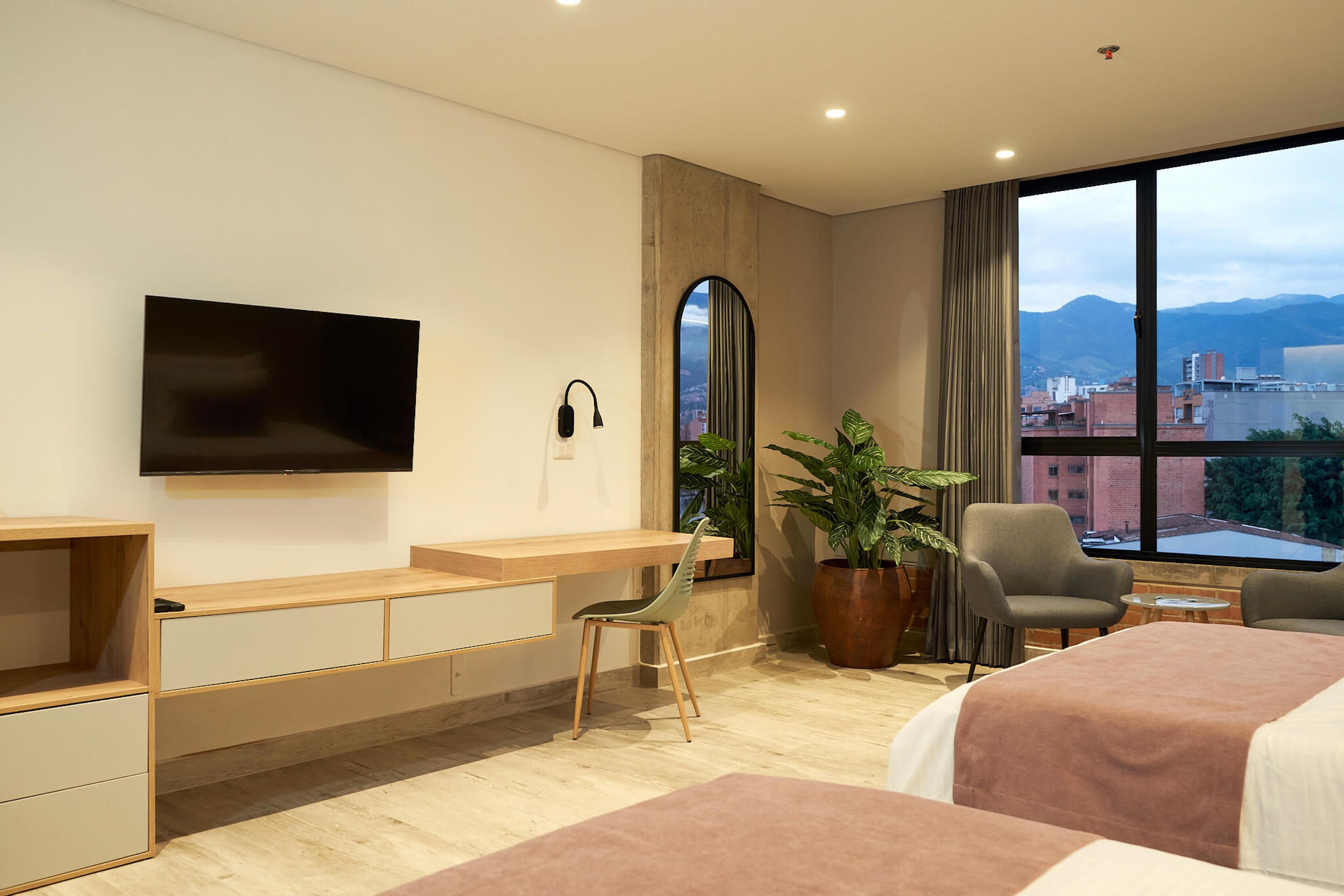 A modern hotel room with a large window, a flat-screen TV, a desk, two chairs, a bed, and a potted plant.