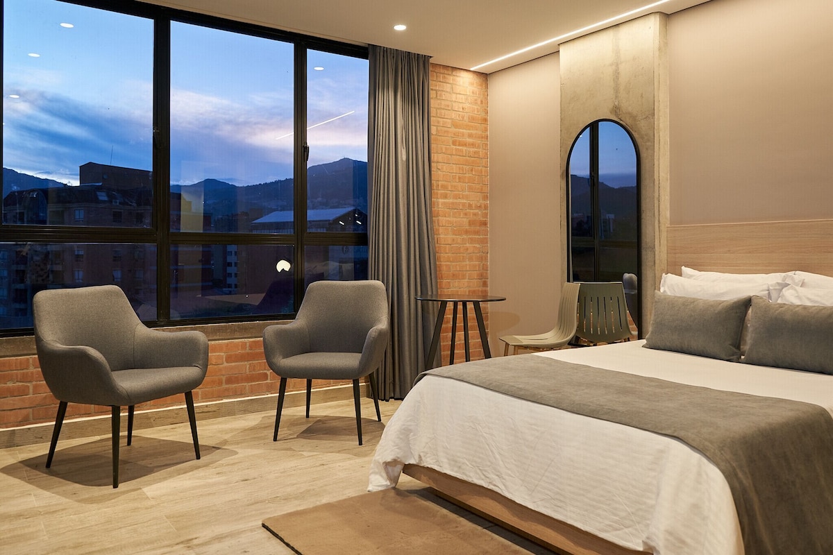 A hotel room with a large bed, two armchairs, a small table, and a view of the cityscape through large windows.